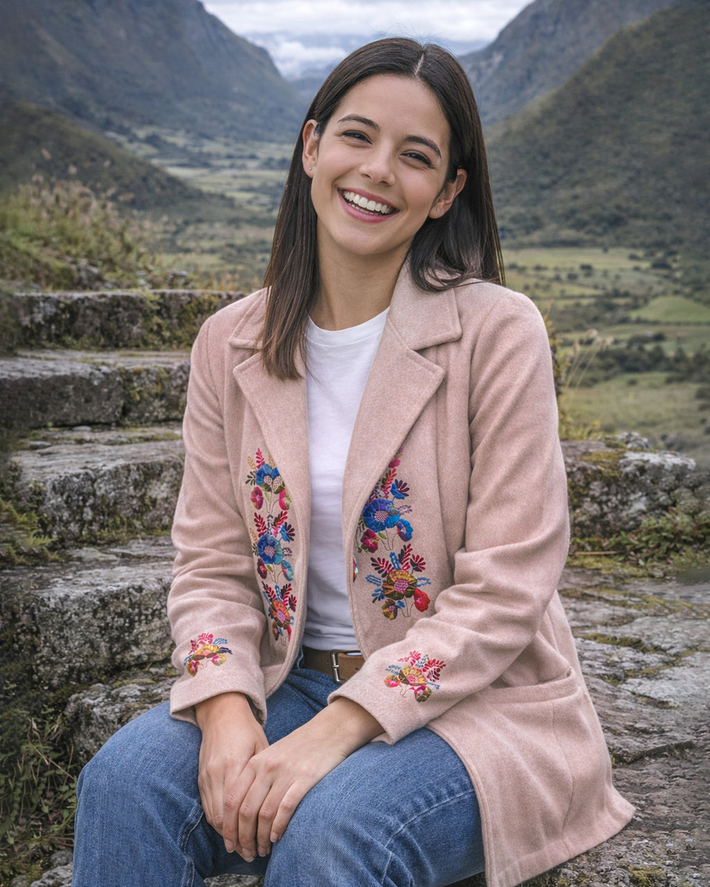 Mujer sentada en unas gradas de piedra con el Abrigo Jardín rosa claro y sus flores bordadas, con montañas al fondo