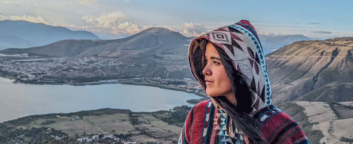 Mujer con el poncho Cayambe y la capucha puesta, frente a una laguna de la sierra