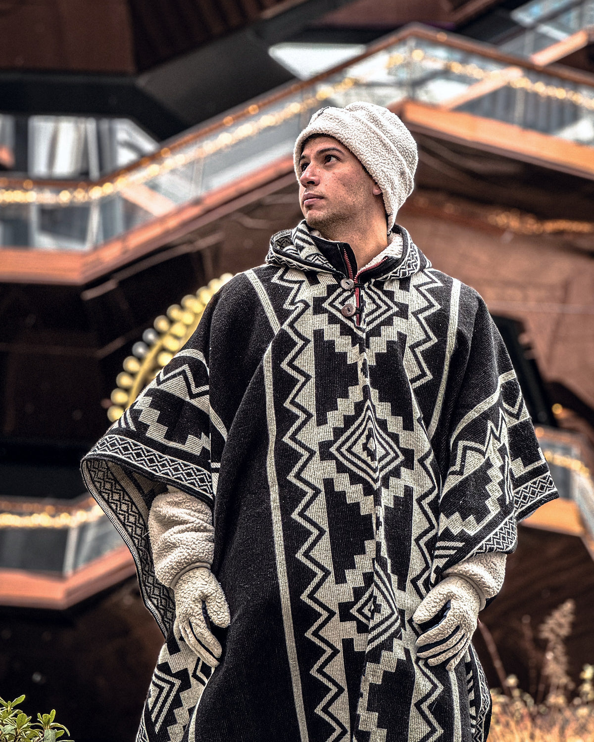 Hombre con gorro de lana y guantes con el poncho Chimborazo negro y crema, en un edificio con luces cálidas