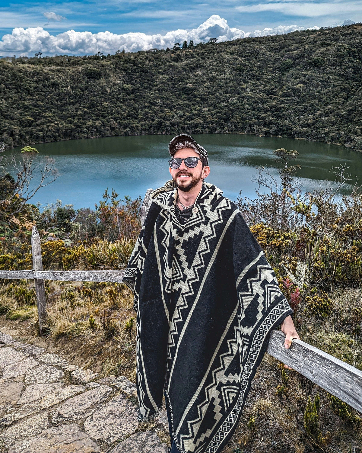 Hombre con gorra y gafas con el poncho Chimborazo negro y crema, junto a una laguna rodeada de cerros