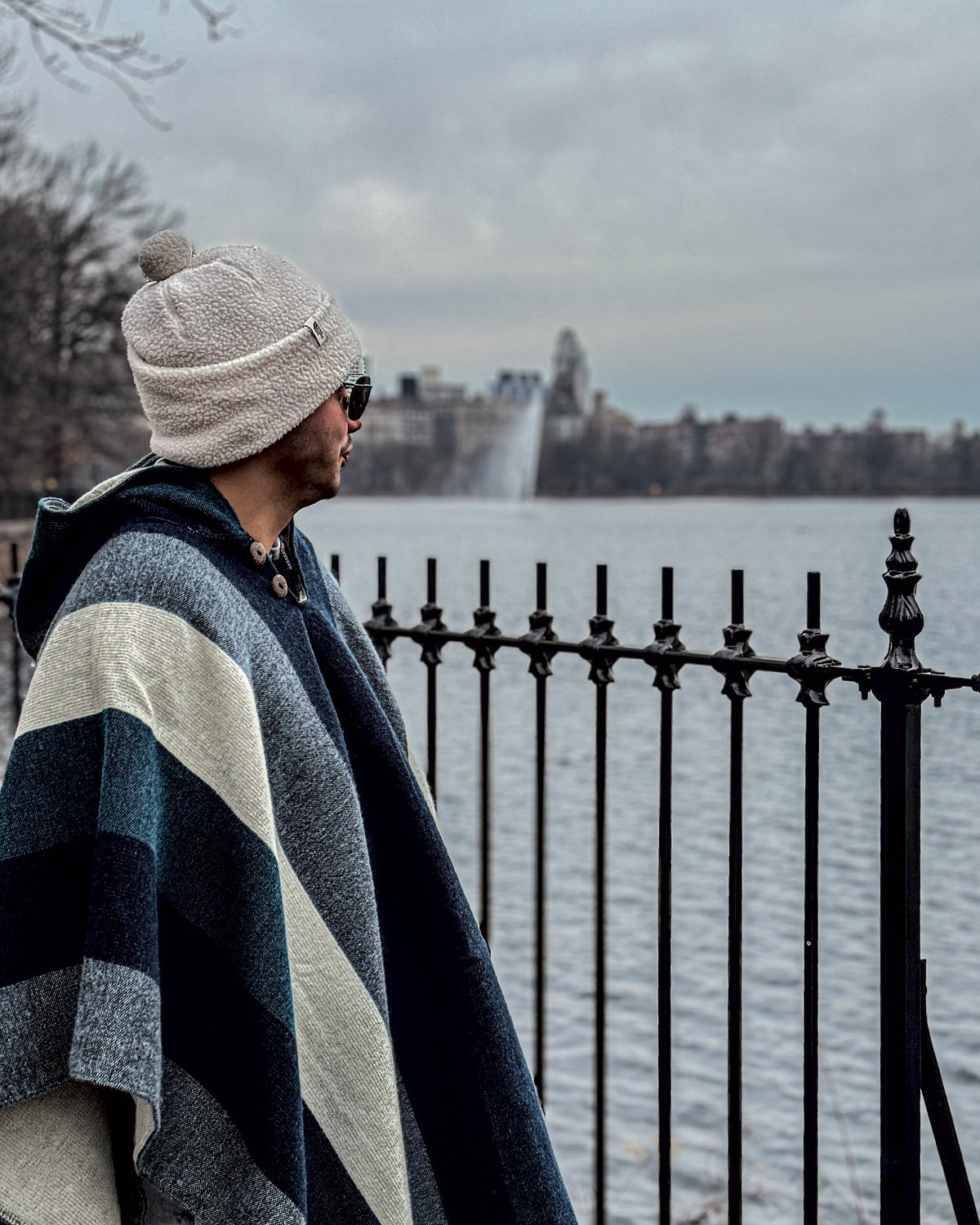 Hombre con gorro de lana y gafas con el poncho Cuicocha, de perfil junto a una baranda frente a un lago