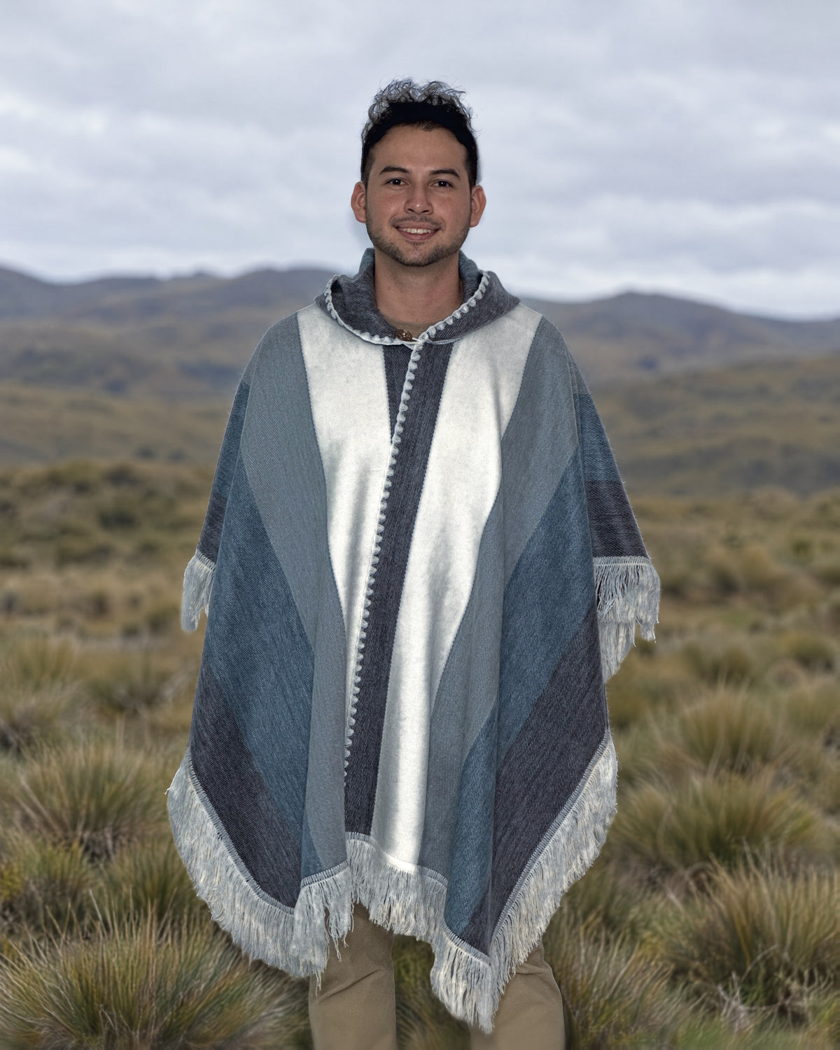 Hombre de pie con el poncho Espíritu Cielo Andino en un campo de pajonal con cerros al fondo