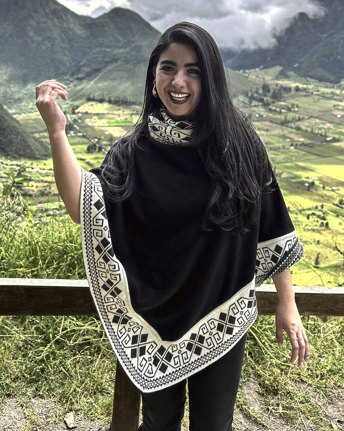 Mujer sonriendo de medio cuerpo con el poncho Lluvia Nocturna negro de borde crema, frente a un valle verde
