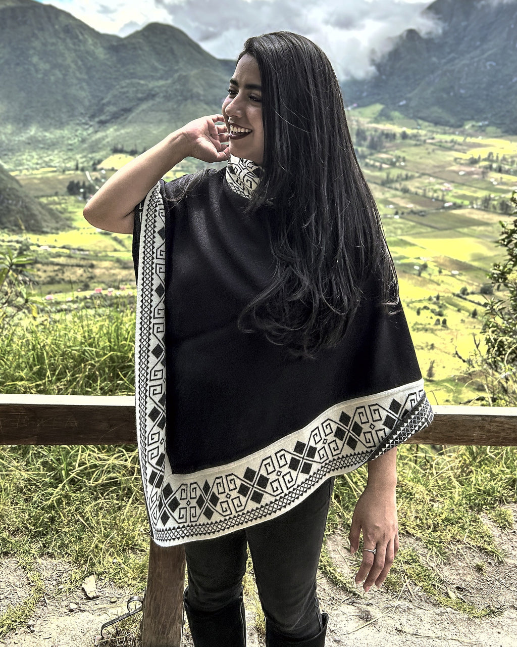 Mujer de tres cuartos con el poncho Lluvia Nocturna negro de borde crema, mirando de lado con montañas al fondo