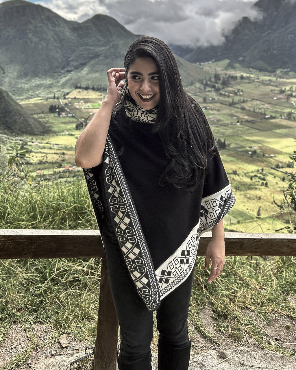 Mujer sonriendo con el poncho Lluvia Nocturna negro y crema, junto a una baranda de madera en un mirador