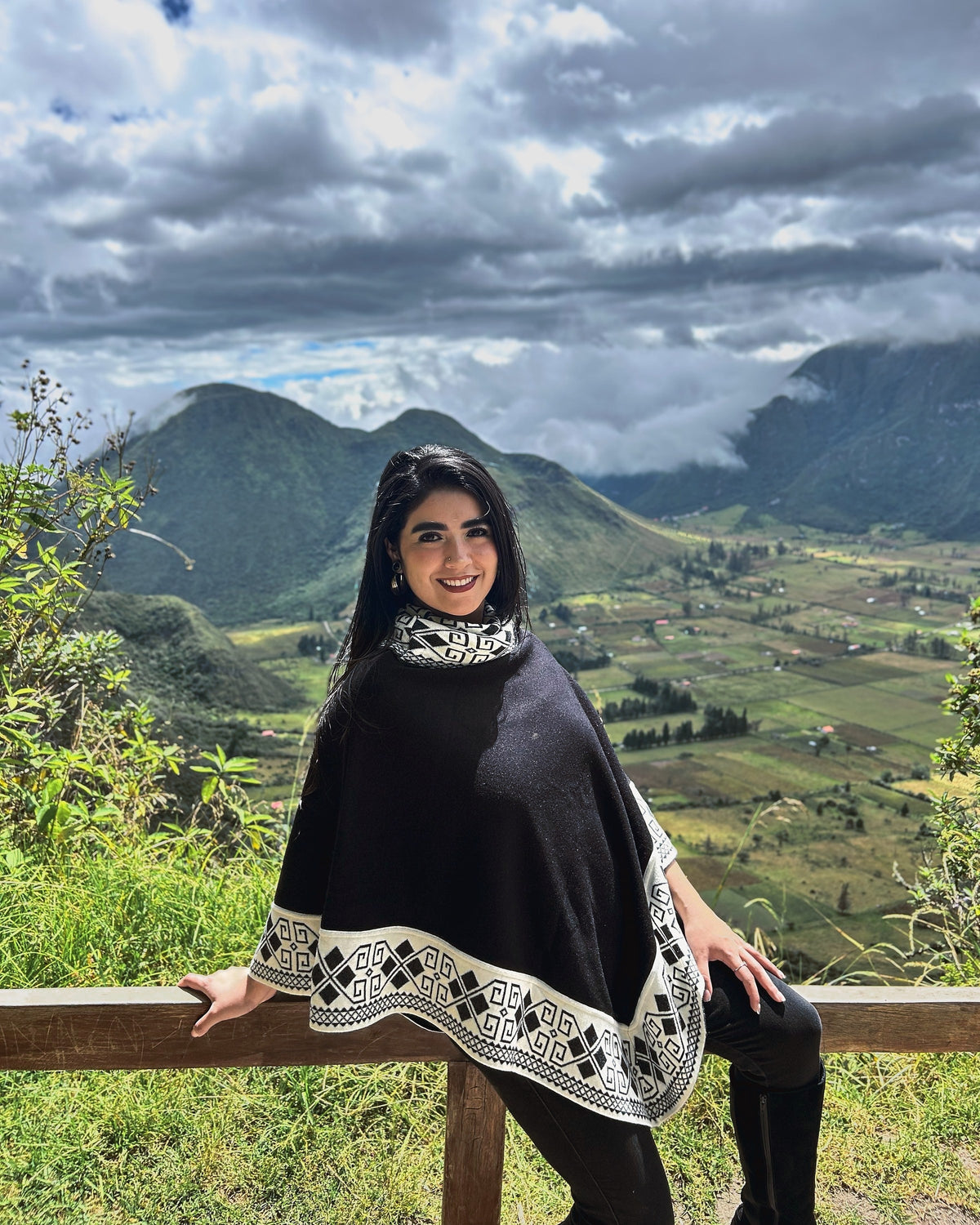 Mujer sentada en una baranda con el poncho Lluvia Nocturna negro y crema, con el valle del Pululahua detrás