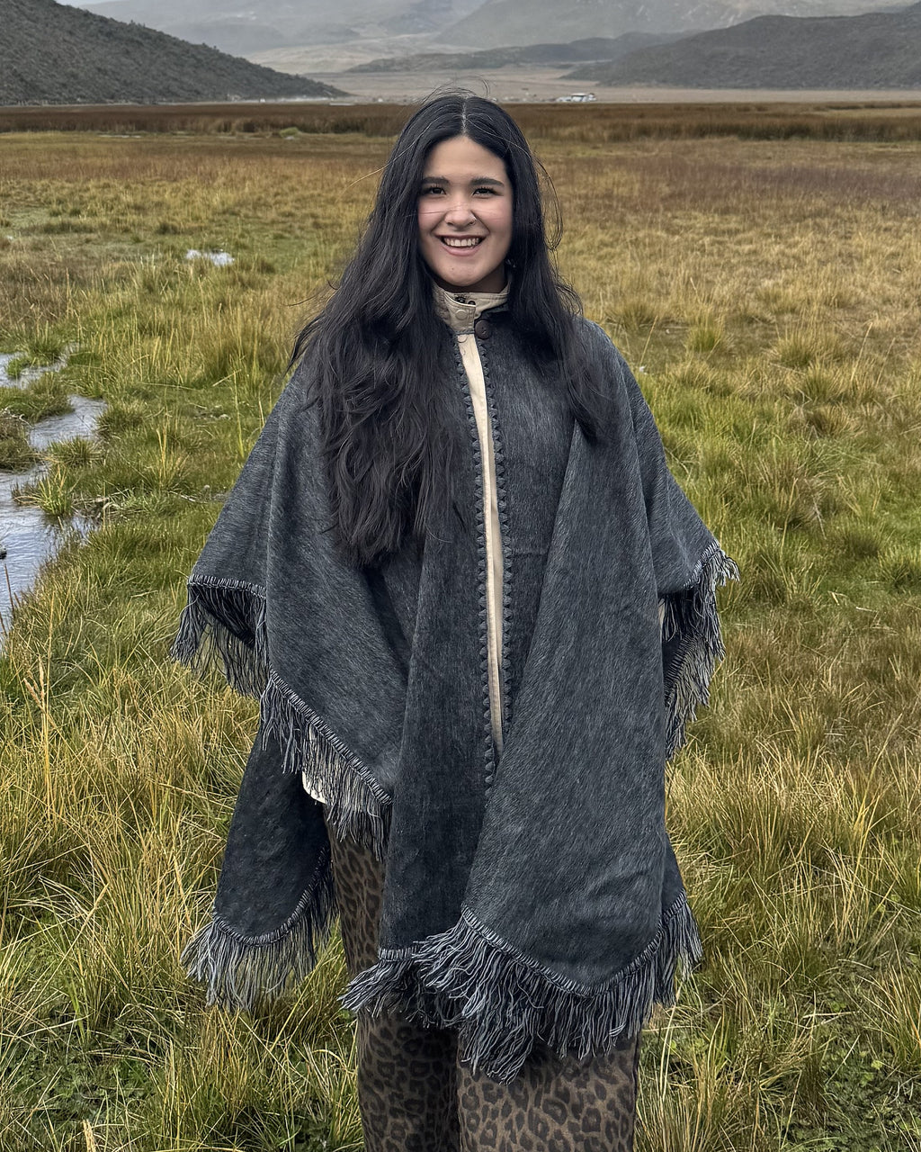 Mujer sonriente con el Poncho Sombra Andina gris abierto al frente y flecos, en un páramo, medio cuerpo