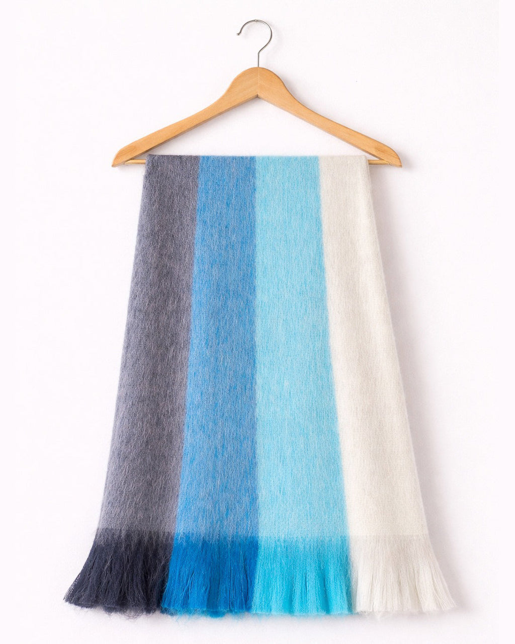 Chal de alpaca Bruma Azul en franjas de gris, azul, celeste y marfil, colgado de una percha de madera