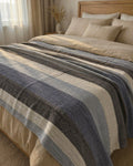Cobija de alpaca Celeste Andino en franjas azules, celestes, crema y gris sobre una cama con almohadas beige