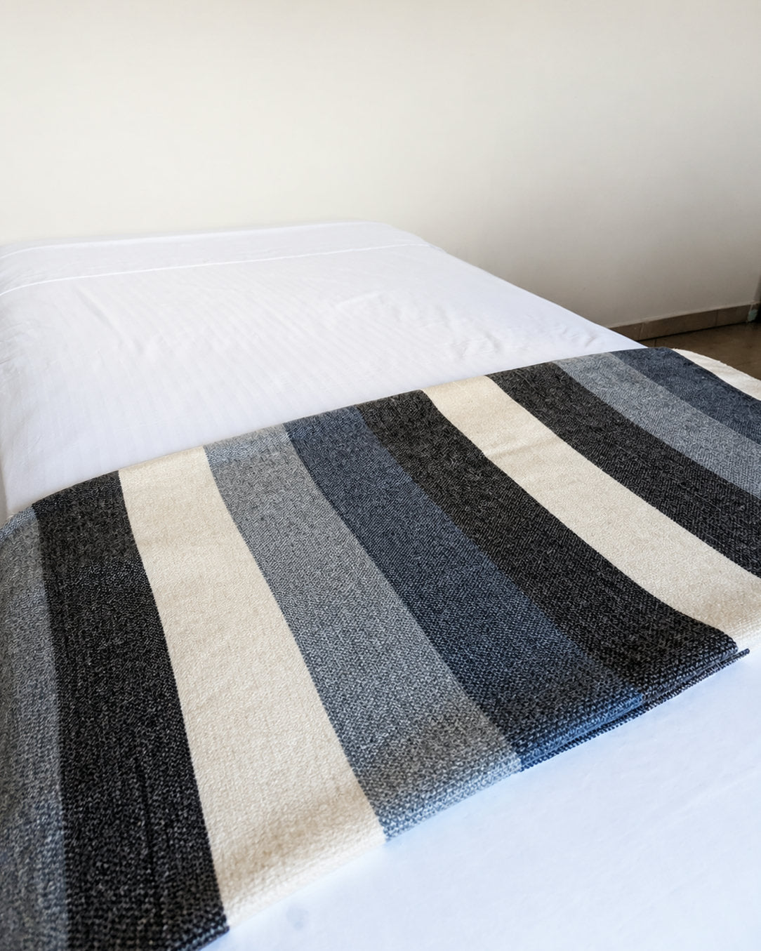 Cobija de alpaca Celeste Andino con franjas azules, crema y grises extendida al pie de una cama blanca