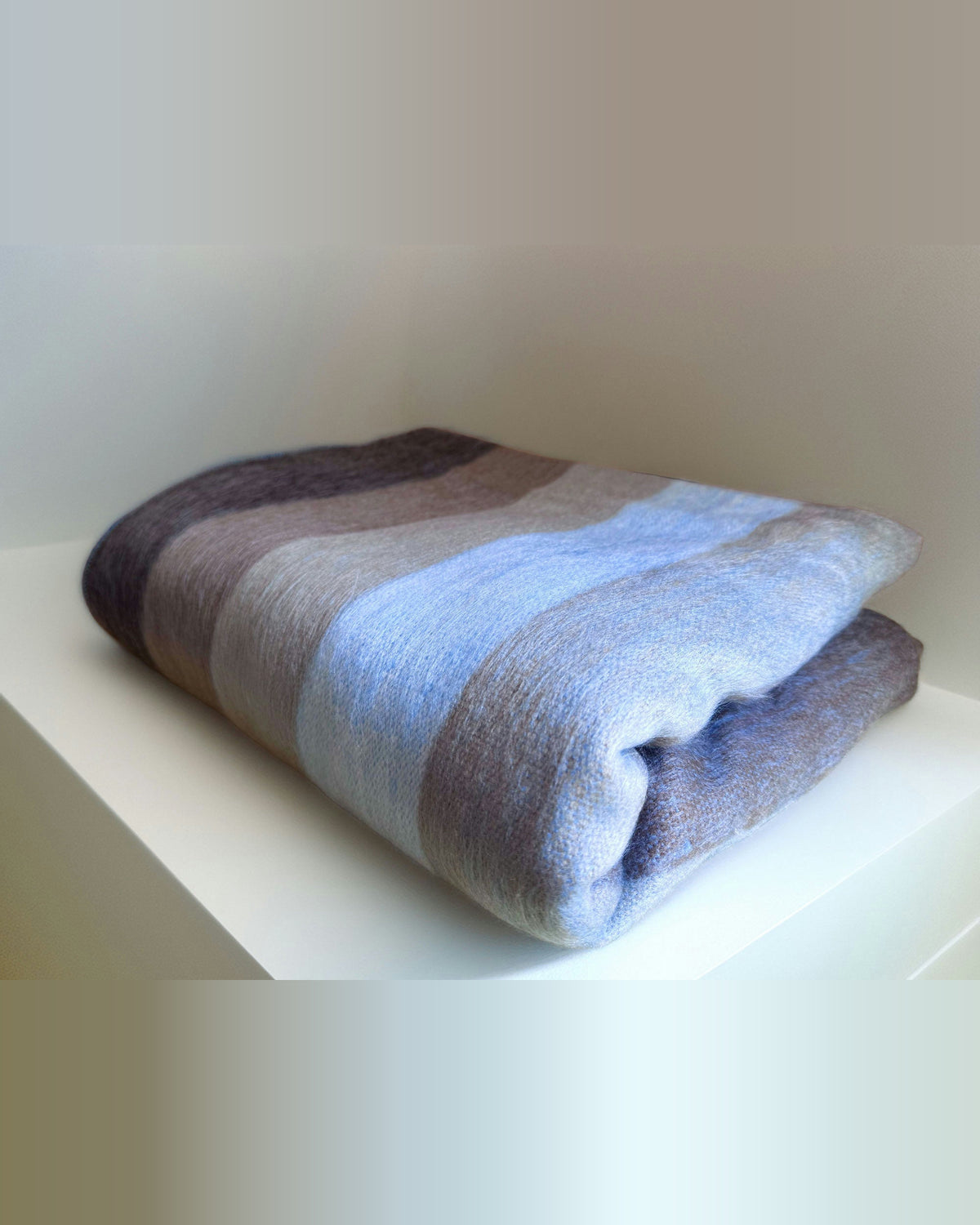 Cobija de alpaca Raíz del Cielo doblada sobre una repisa blanca, con franjas azul claro, lila y café