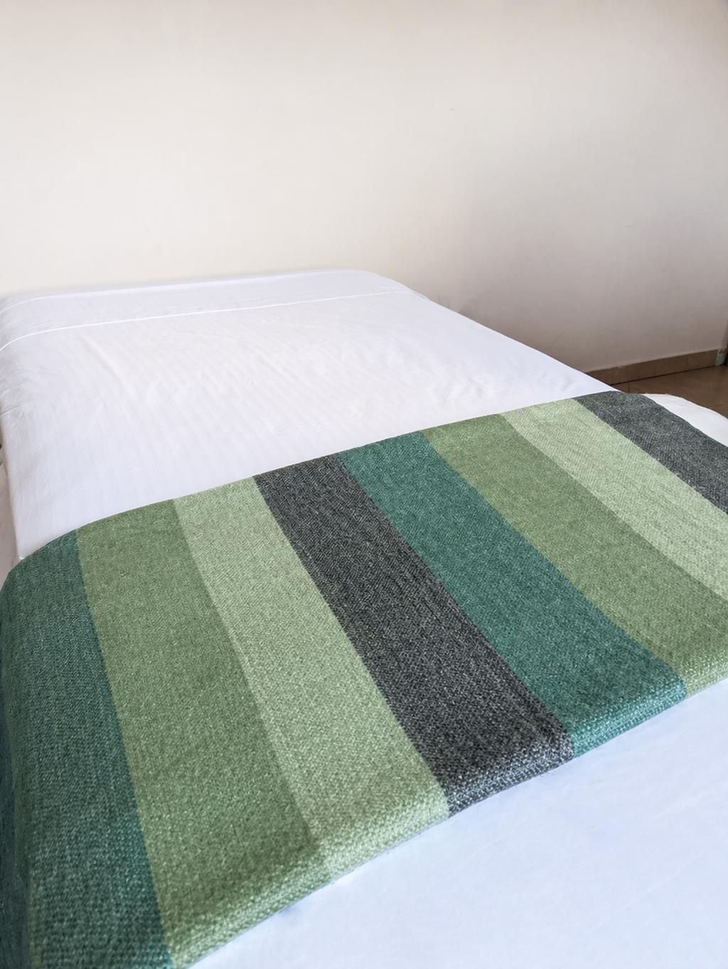 Cobija de alpaca Sendero del Bosque con franjas verde azulado, salvia y gris oscuro al pie de una cama blanca