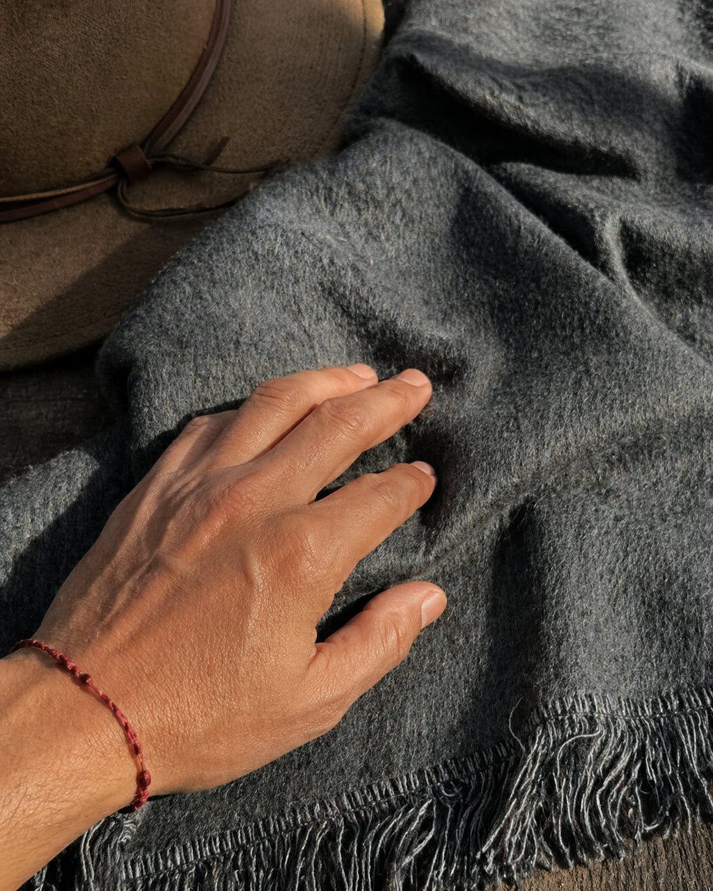 Mano sobre el tejido suave del Poncho Sombra Andina gris, con un sombrero detrás